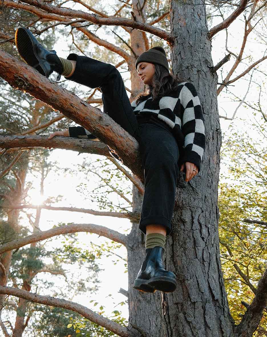 Co-founder of RAWTY Sophia Stöhr sitting in a tree wearing a beanie, a checkered pullover, black pants and black shoes in soft sunlight.