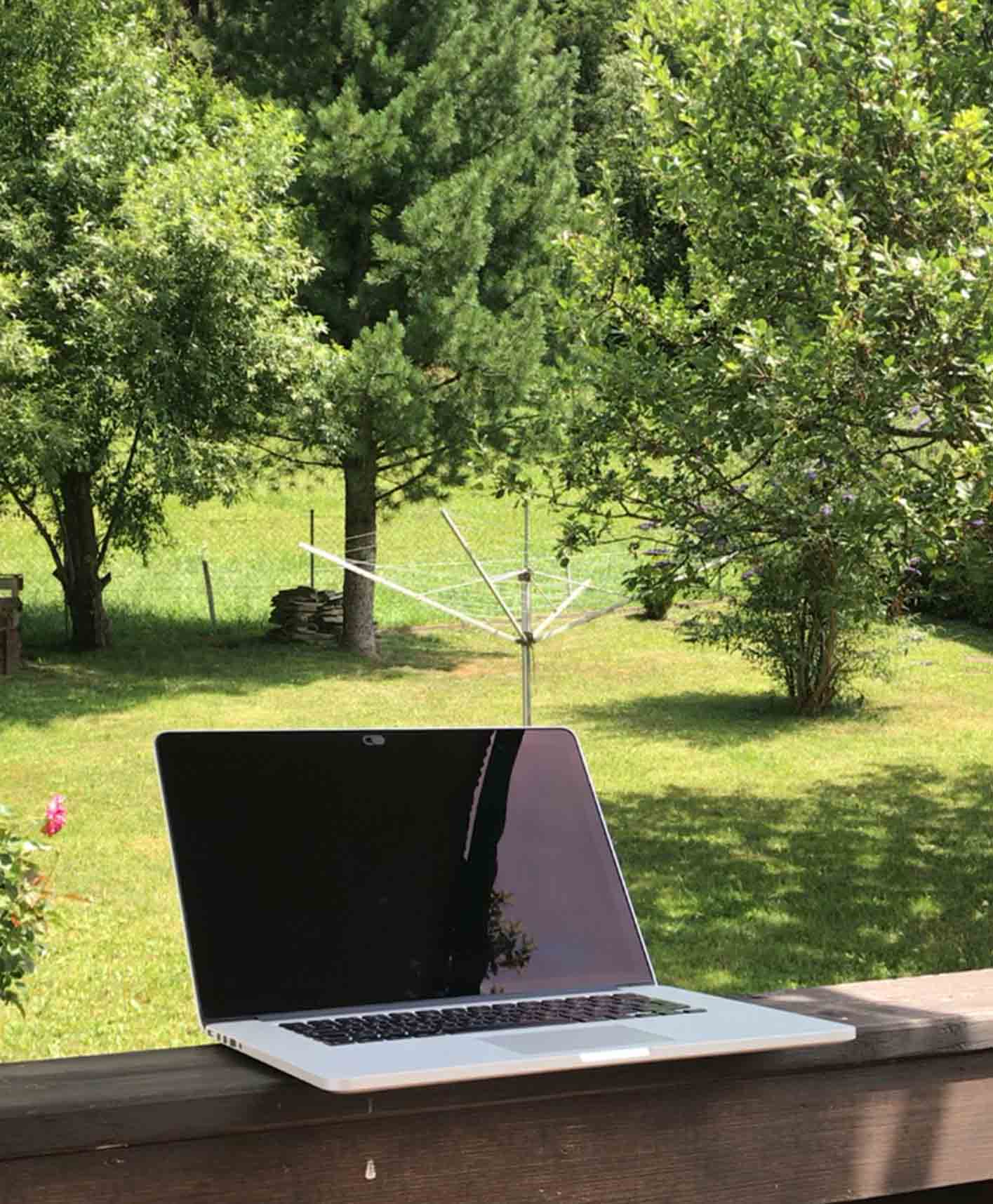 An open laptop standing on a balcony overlooking a green garden scene with trees and grass in Austria.