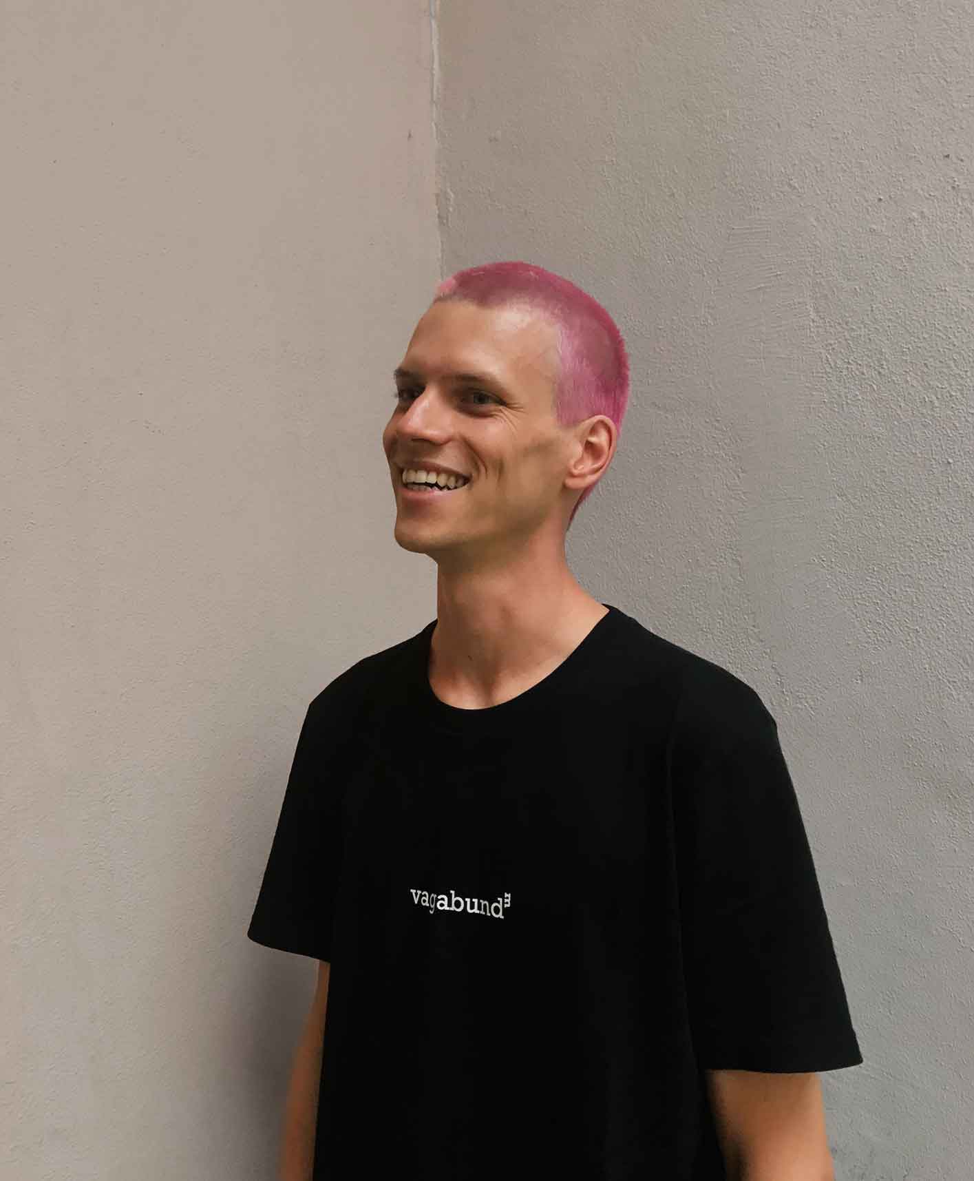 Portrait of Christian Leban with short pastel pink hair wearing a black t-shirt standing in front of a wall.