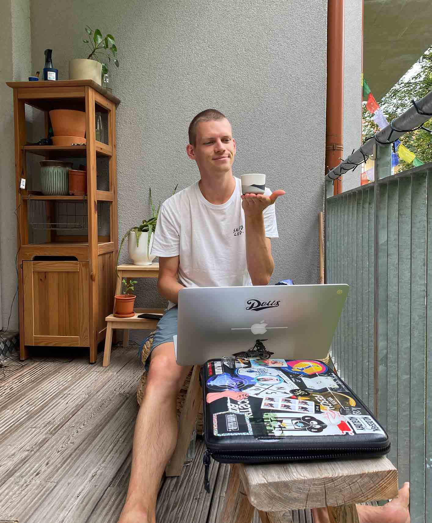 Christian Leban sitting on a balcony working on his laptop and holding a cup of coffee looking happy.