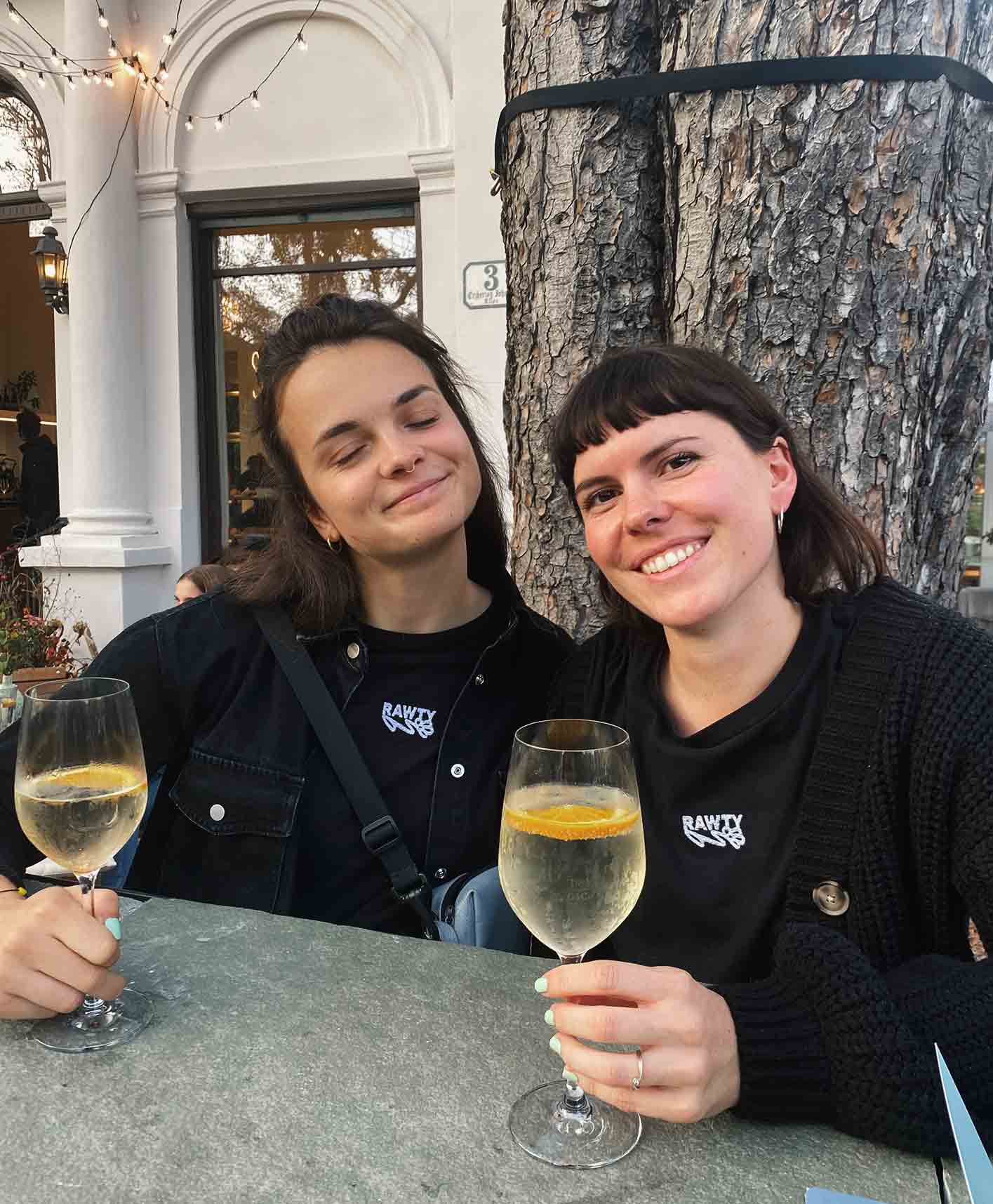 Sophia Stöhr and Tessa Huber sitting outside having a drink, smiling and wearing black RAWTY t-shirts.