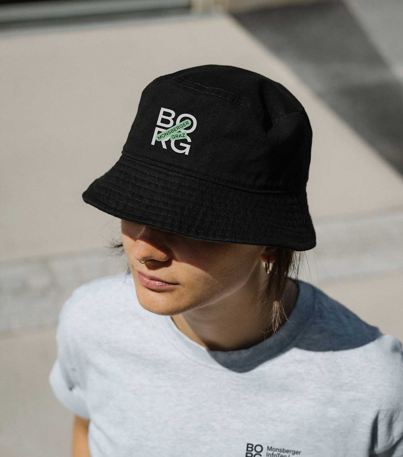 A female student wearing a black bucket hat with the BORG Monsberger logo and a grey t-shirt with the logo in bright sunlight shot from slightly above.