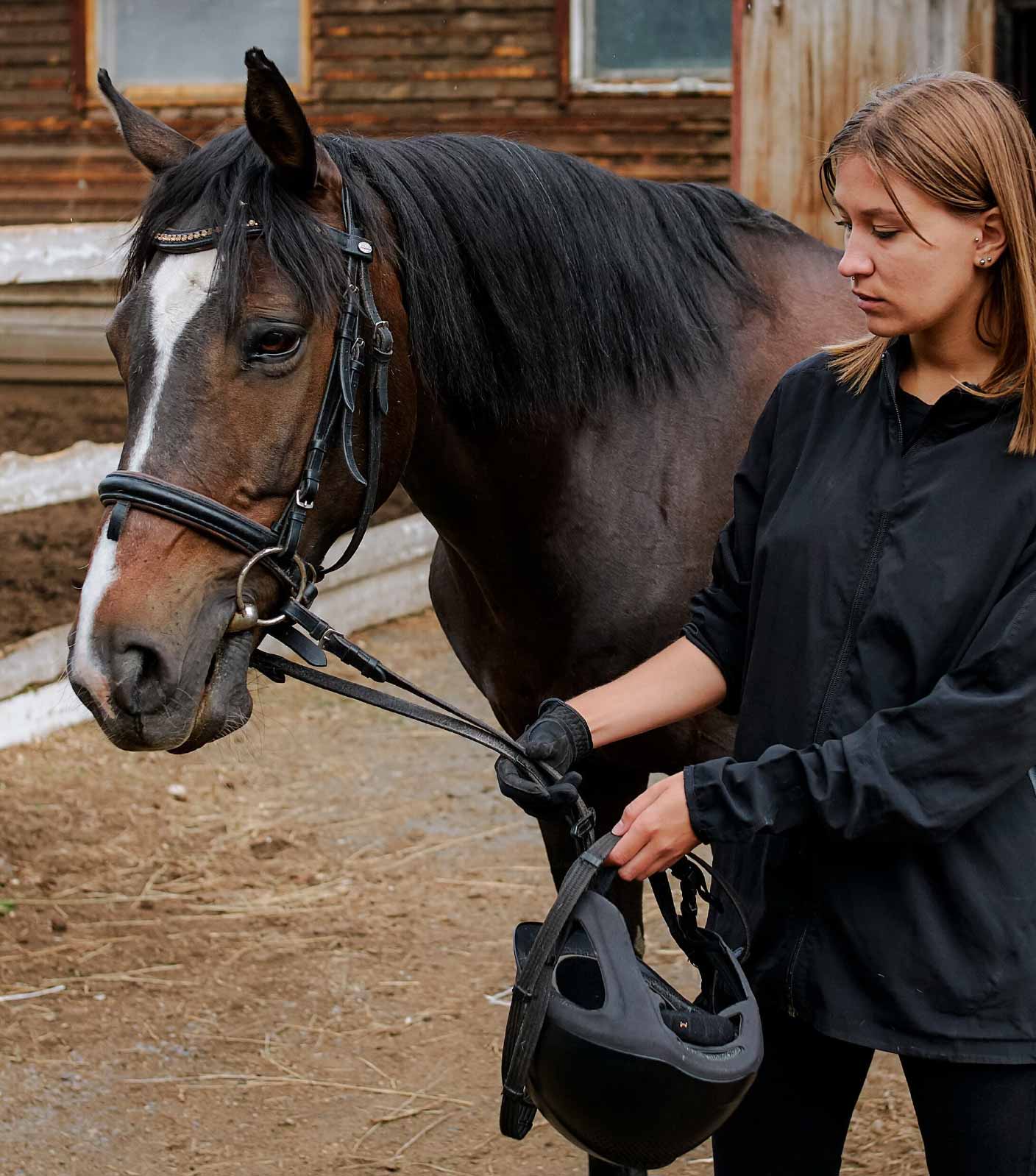 A female student dressed in black standing next to a dark brown horse holding the reins and looking at the horse.