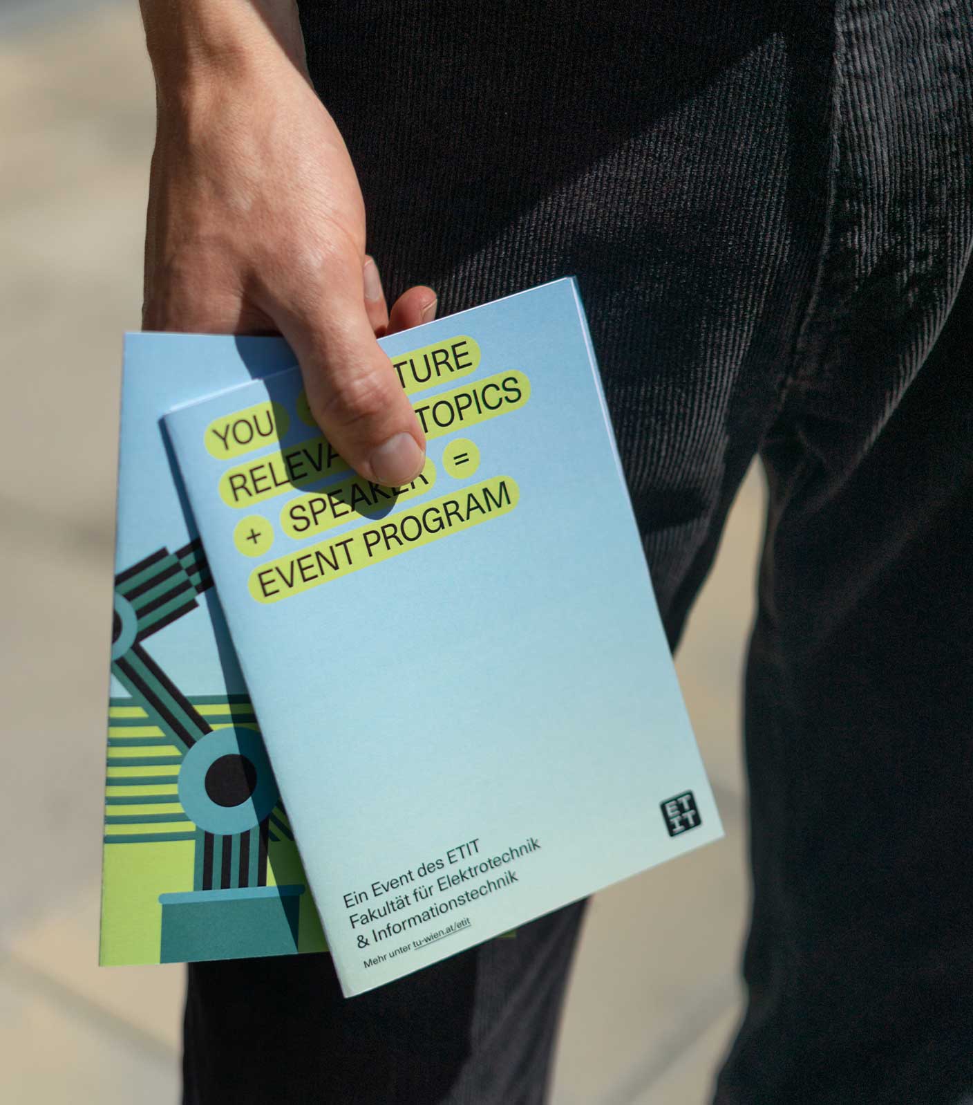 A photo of a person holding two brochures of the ETIT near their paints showing a cover with an equation title and the logo and the other brochure cover showing a vector illustration of a robotic arm, all in the neon green-yellow, green, light-blue and black color scheme.
