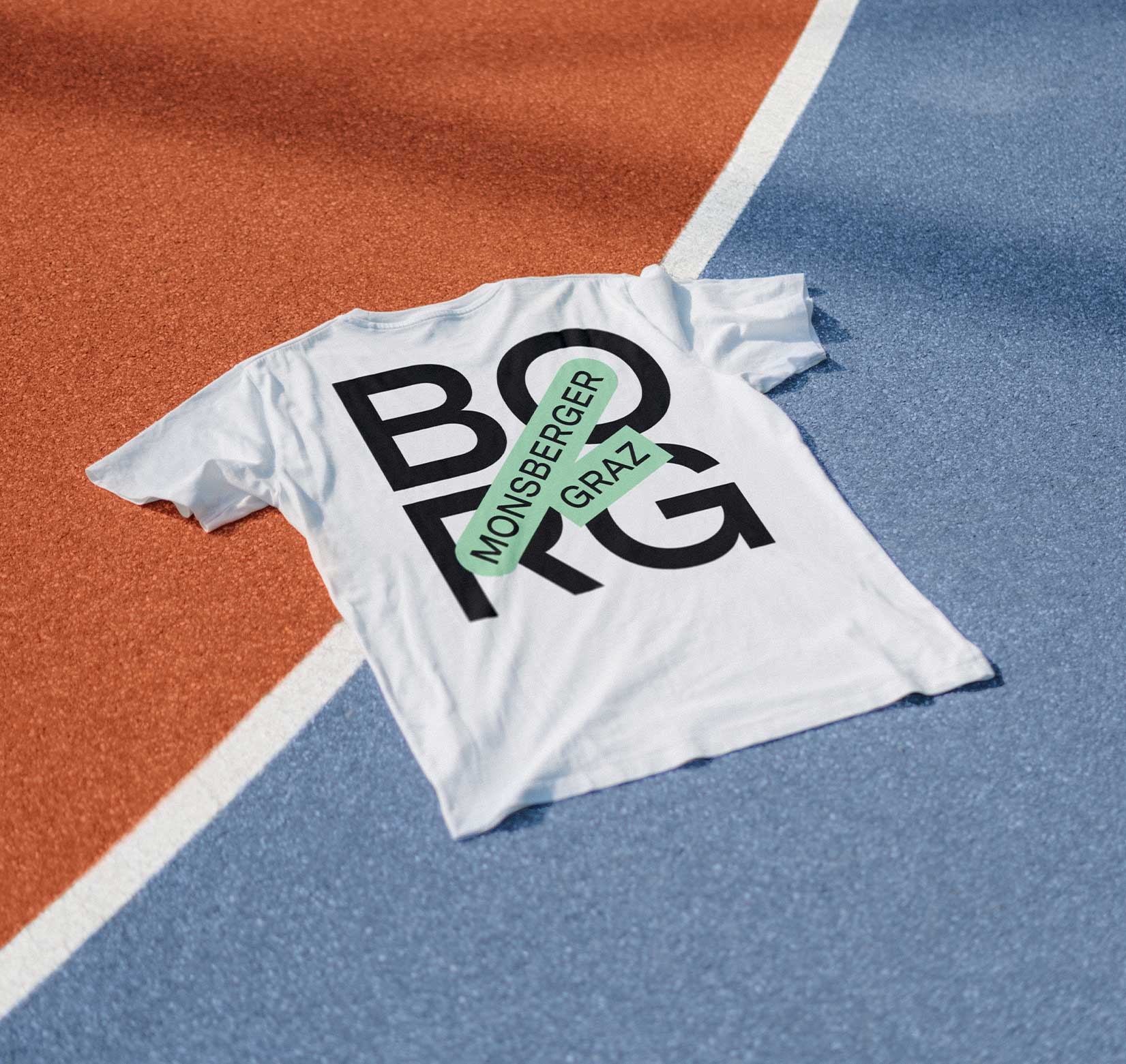A white t-shirt with the BORG Monsberger logo on the back, lying flat on the ground at a blue and orange basketball court.