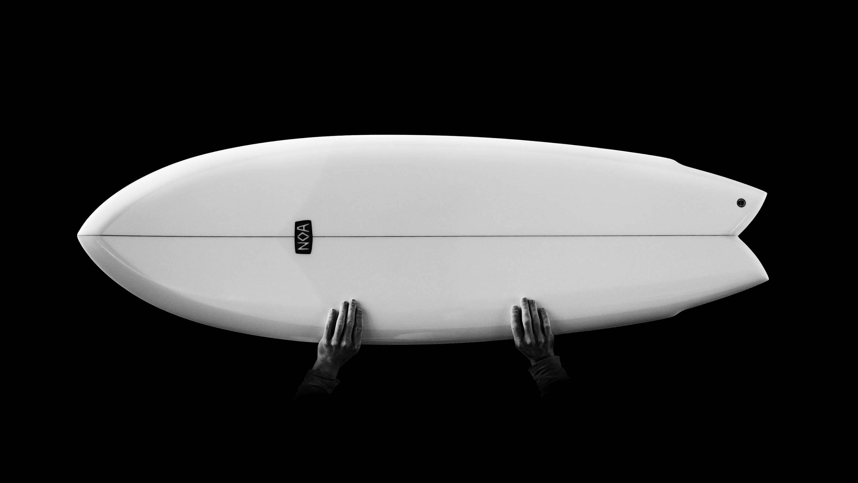 Two hands holding a white surfboard in fish shape horizontally in the air on a black background in harsh light that highlights the beautiful curves of the surfboard.