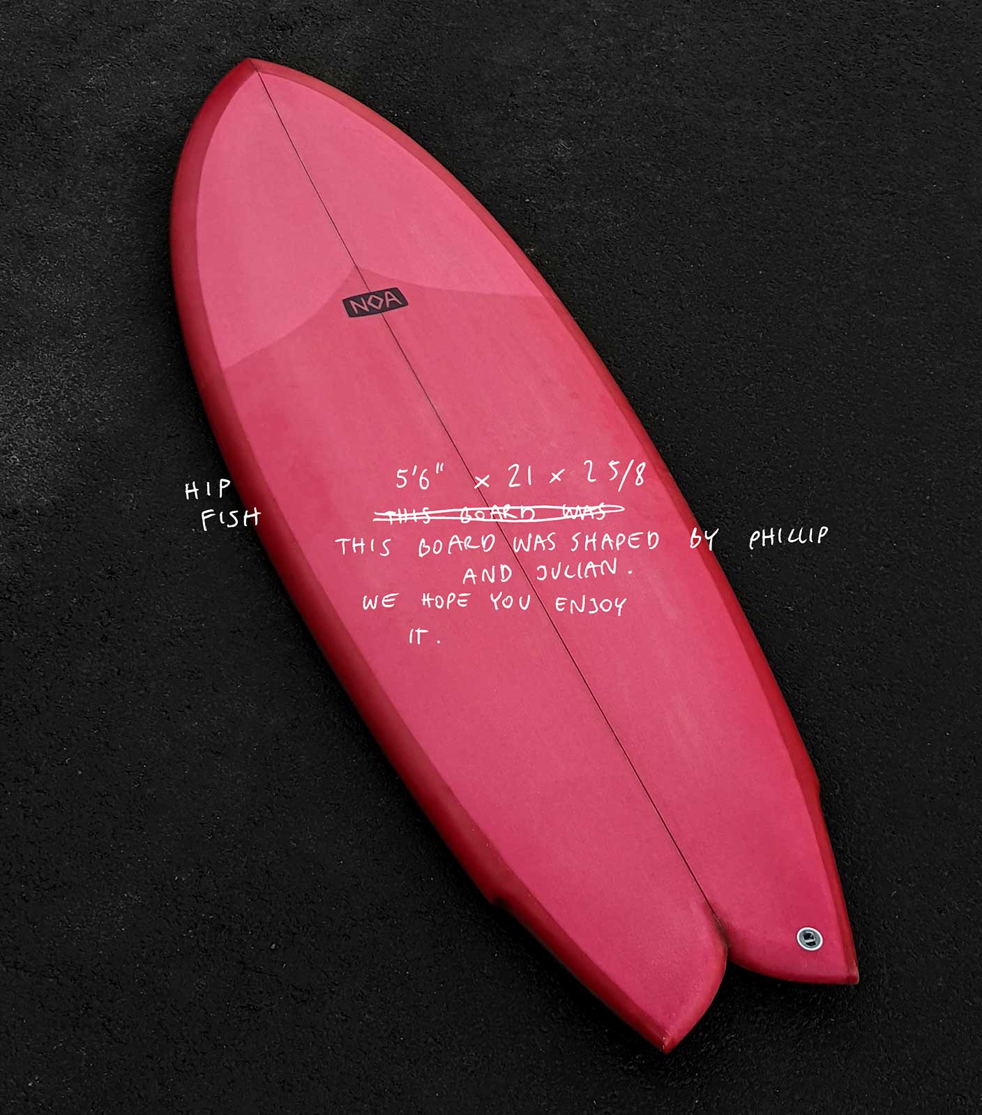 A red surfboard in fish shape lying on black asphalt floor with white descriptive text written on the photo that says 