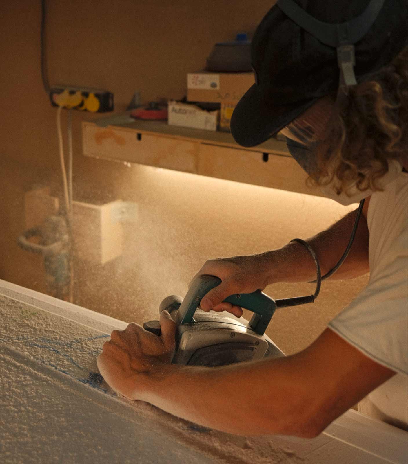 Surfboard shaper Phillip Tritthart shaping a surfboard wearing a black cap and a white t-shirt in the workshop with dust flying around.