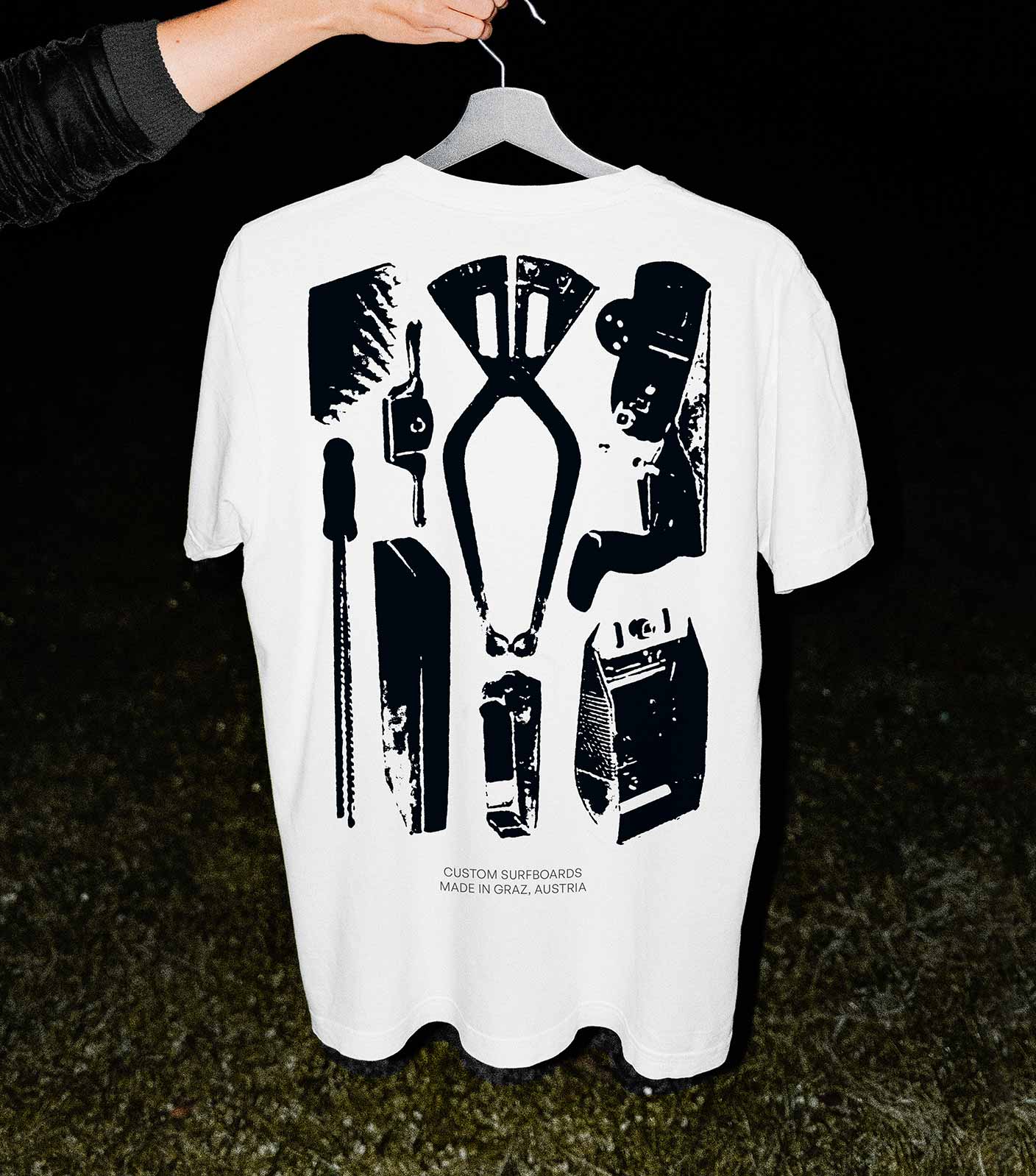 A hand holding a black hanger with a white t-shirt on it that has high contrast black graphics of shape tools as a backprint. The photo is shot with flash in the night where only the t-shirt and the hand and a little of the grass floor is visible.