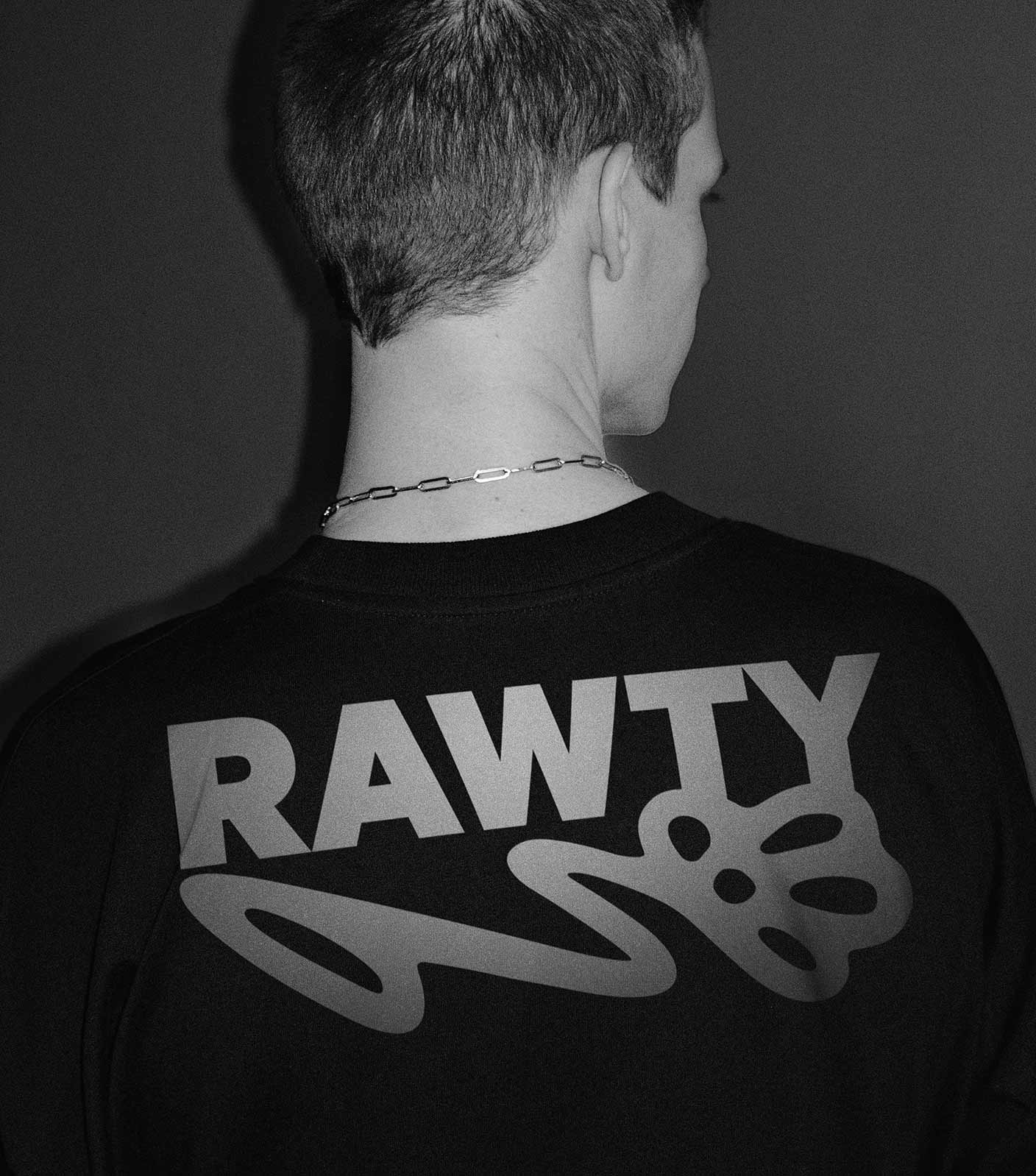 A black and white photo of person standing with the back to the camera wearing a black t-shirt with the white RAWTY logo.